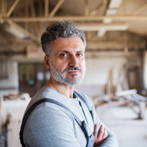 Man working in a carpentry shop looks directly at the camera, with a serious expression on his face.