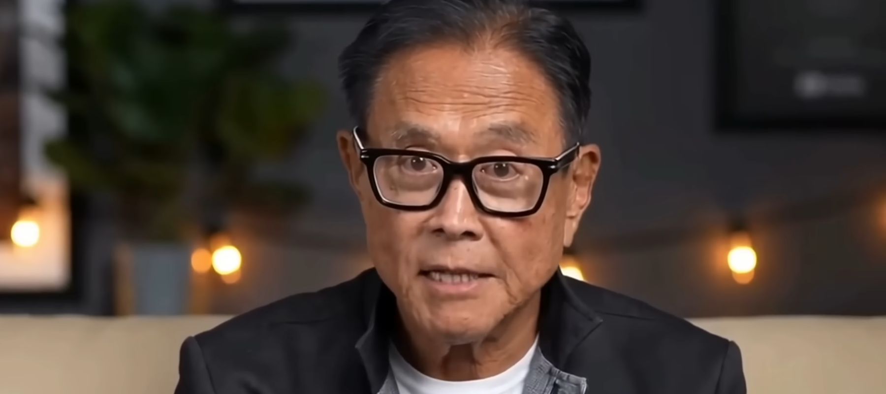 Robert Kiyosaki addresses the viewer empathetically in a black jacket and white shirt.