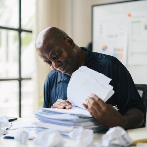 An adult man reviewing papers at a desk surrounded by documents, crumpled papers, and office supplies, creating a busy and messy workspace.