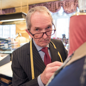 Man looks up at camera with unimpressed look, measuring tape dangles around his neck, while he adjusts a suit on a mannequin.
