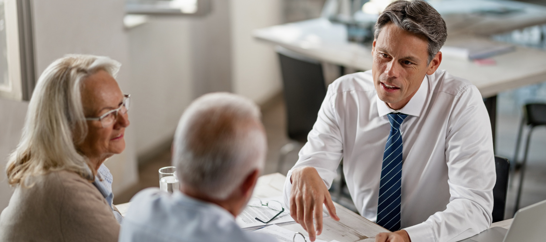 A mature couple is meeting with a financial advisor in a bright office. The advisor is gesturing with his hand while looking at the couple.