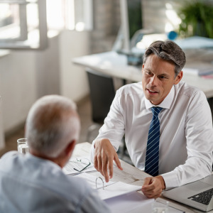 A mature couple is meeting with a financial advisor in a bright office. The advisor is gesturing with his hand while looking at the couple.