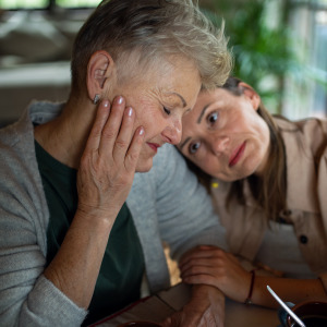 An adult woman with long brown hair rests her head affectionately on a senior woman with short gray hair.