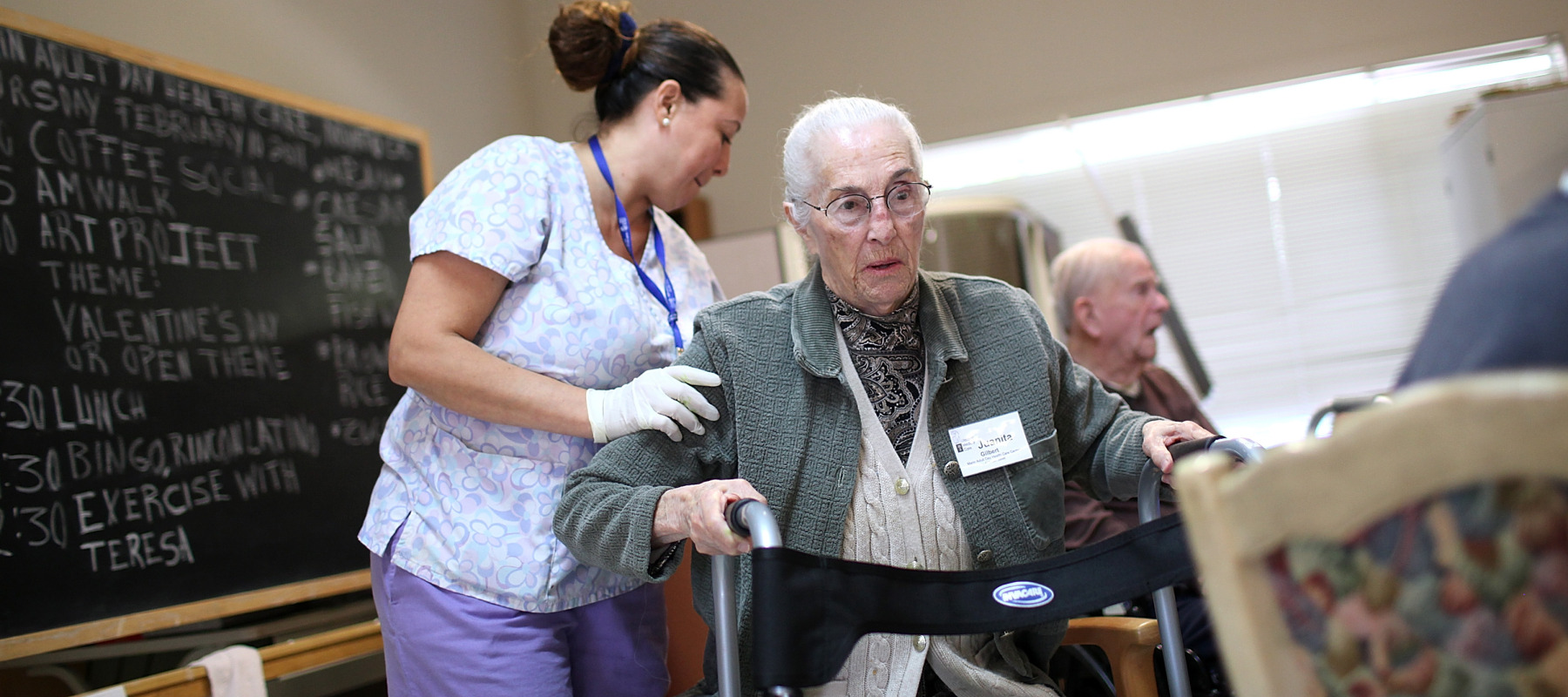 Long-term care nurse helps elderly woman walk inside a building.