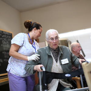 Long-term care nurse helps elderly woman walk inside a building.