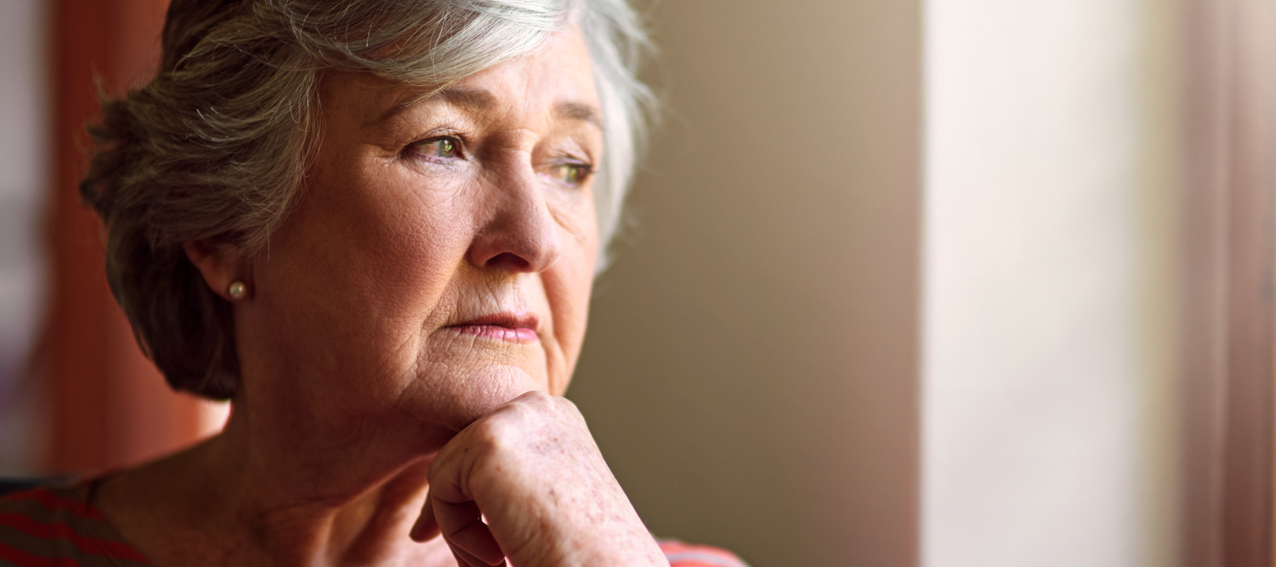 A senior woman with short, wavy grey hair sits indoors in a striped top, gazing out a window.