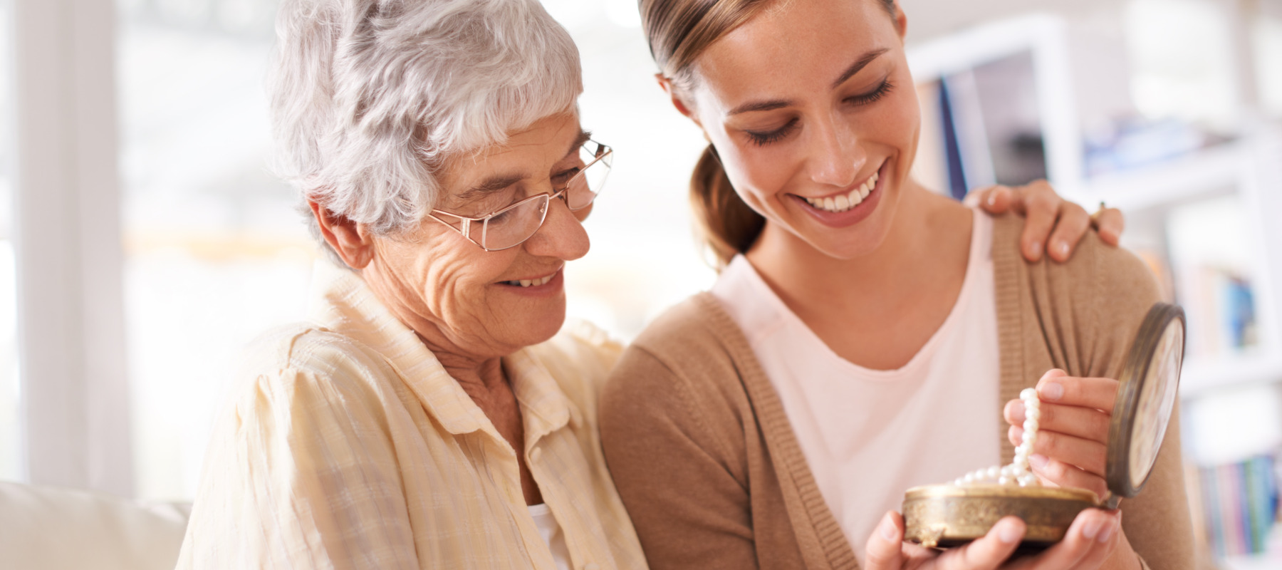 An elderly woman and a younger woman look happily at a piece of jewelry together.
