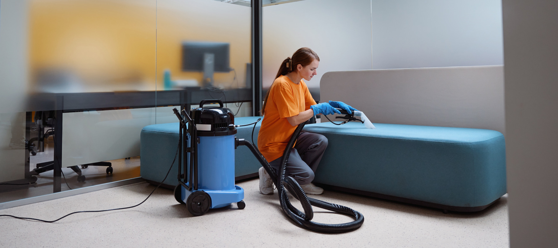 Woman in bright orange t-shirt vacuums couch in an office.