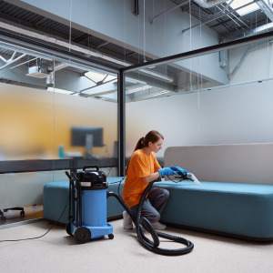 Woman in bright orange t-shirt vacuums couch in an office.