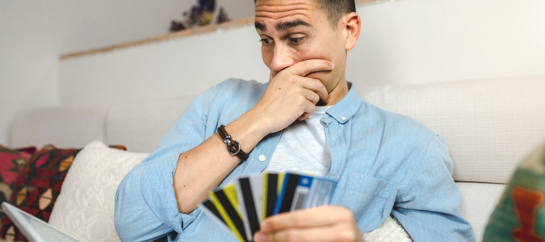 A young adult with short brown hair sits indoors with a tablet and several credit cards in hand, looking concerned.