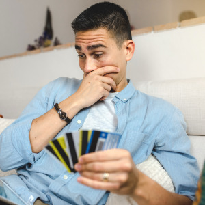 A young adult with short brown hair sits indoors with a tablet and several credit cards in hand, looking concerned.