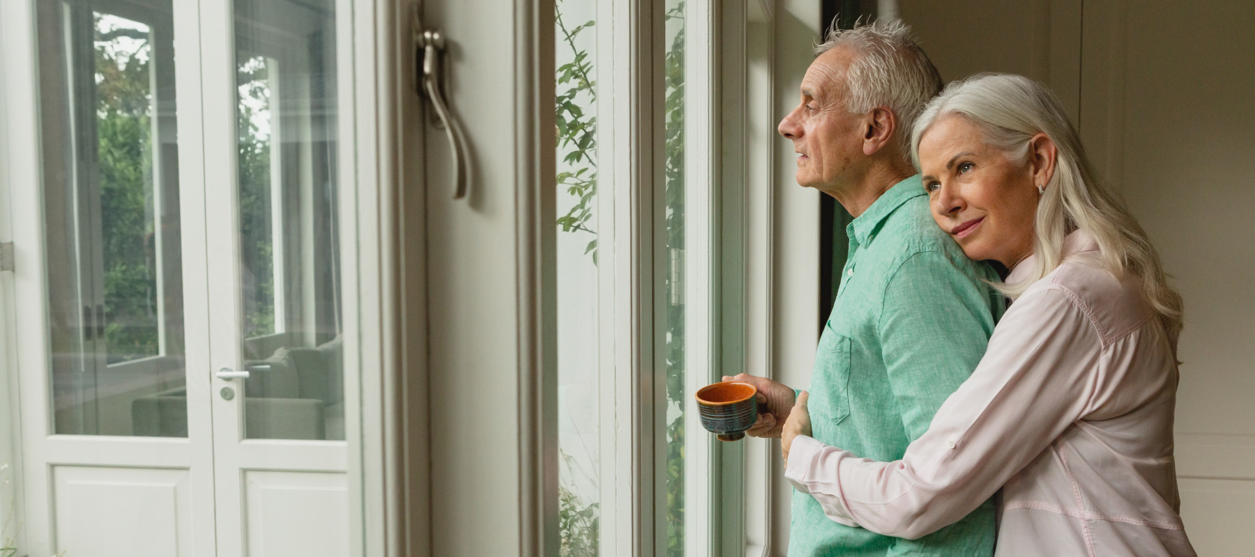 Older couple seen from the side, embracing while looking out the window.