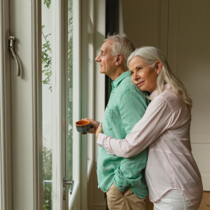 Older couple seen from the side, embracing while looking out the window.
