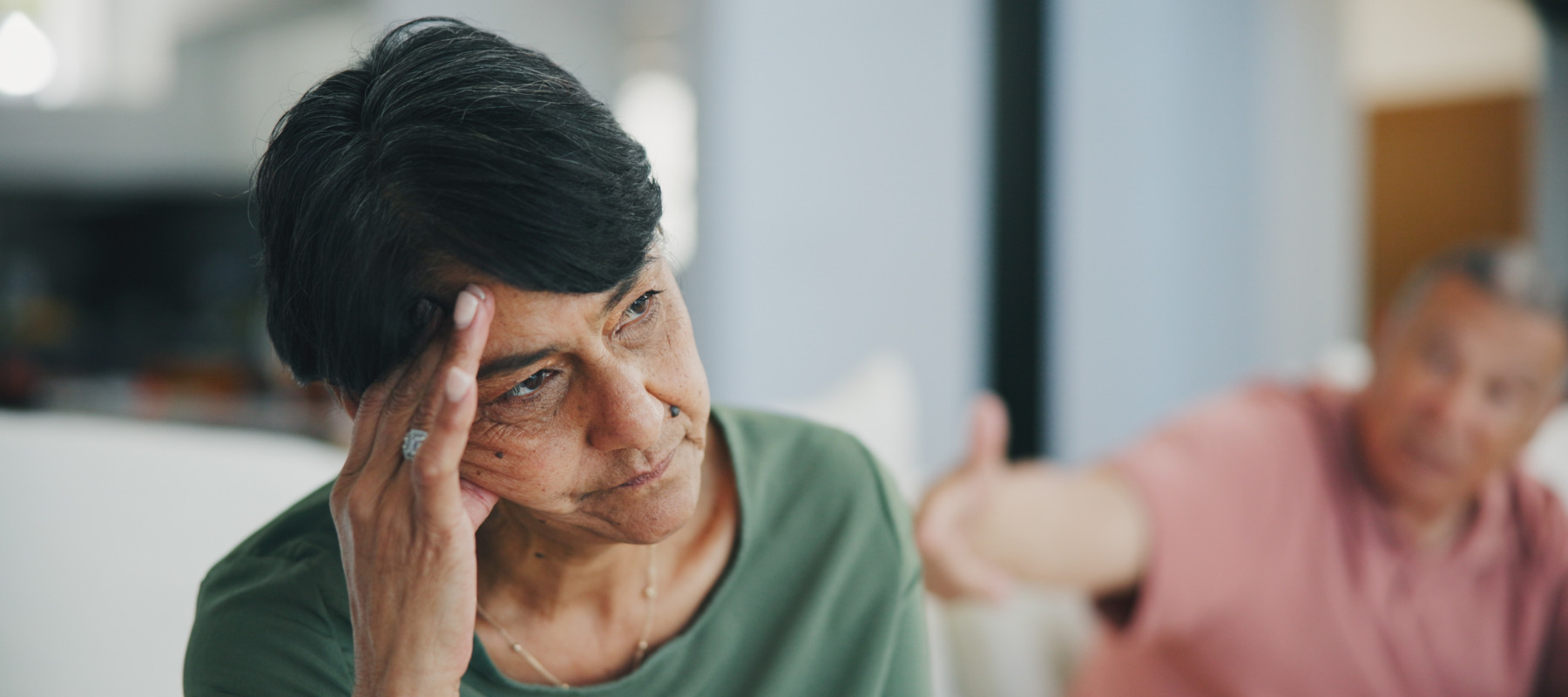 Older woman looks very upset, sitting on the couch, man blurred behind her.
