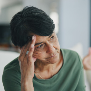 Older woman looks very upset, sitting on the couch, man blurred behind her.