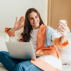 A cheerful young woman with long brown hair sits cross-legged, smiling and waving at her smartphone, as if on a video call.