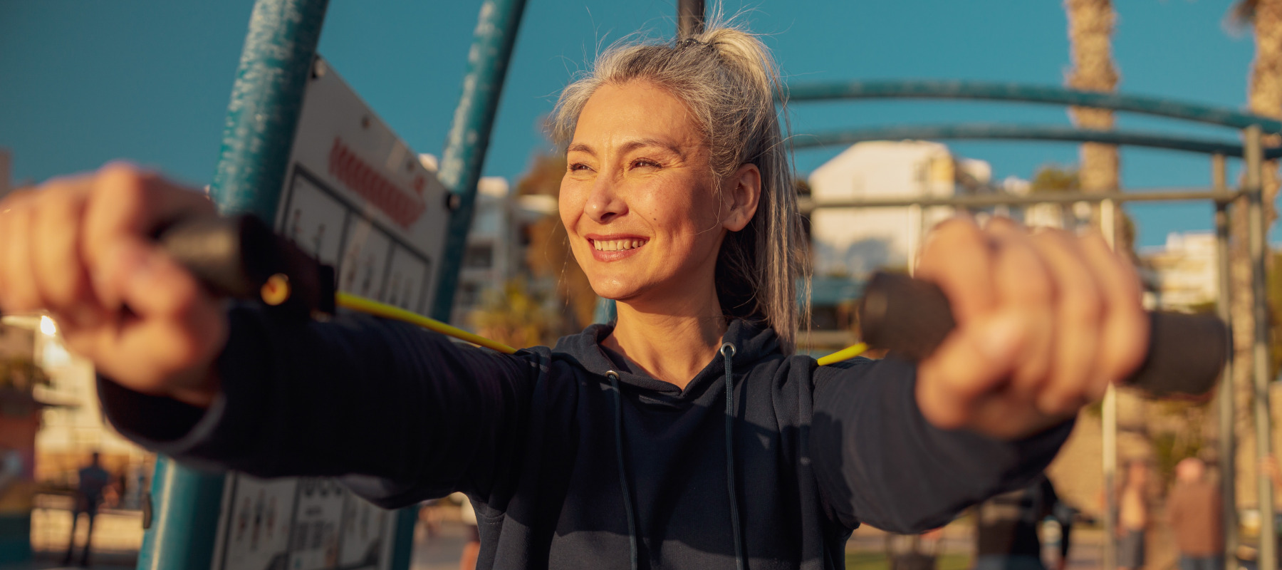 Older woman smiles while stretching exercise bands in front of her at outdoor gym.