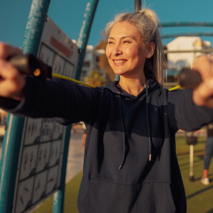 Older woman smiles while stretching exercise bands in front of her at outdoor gym.