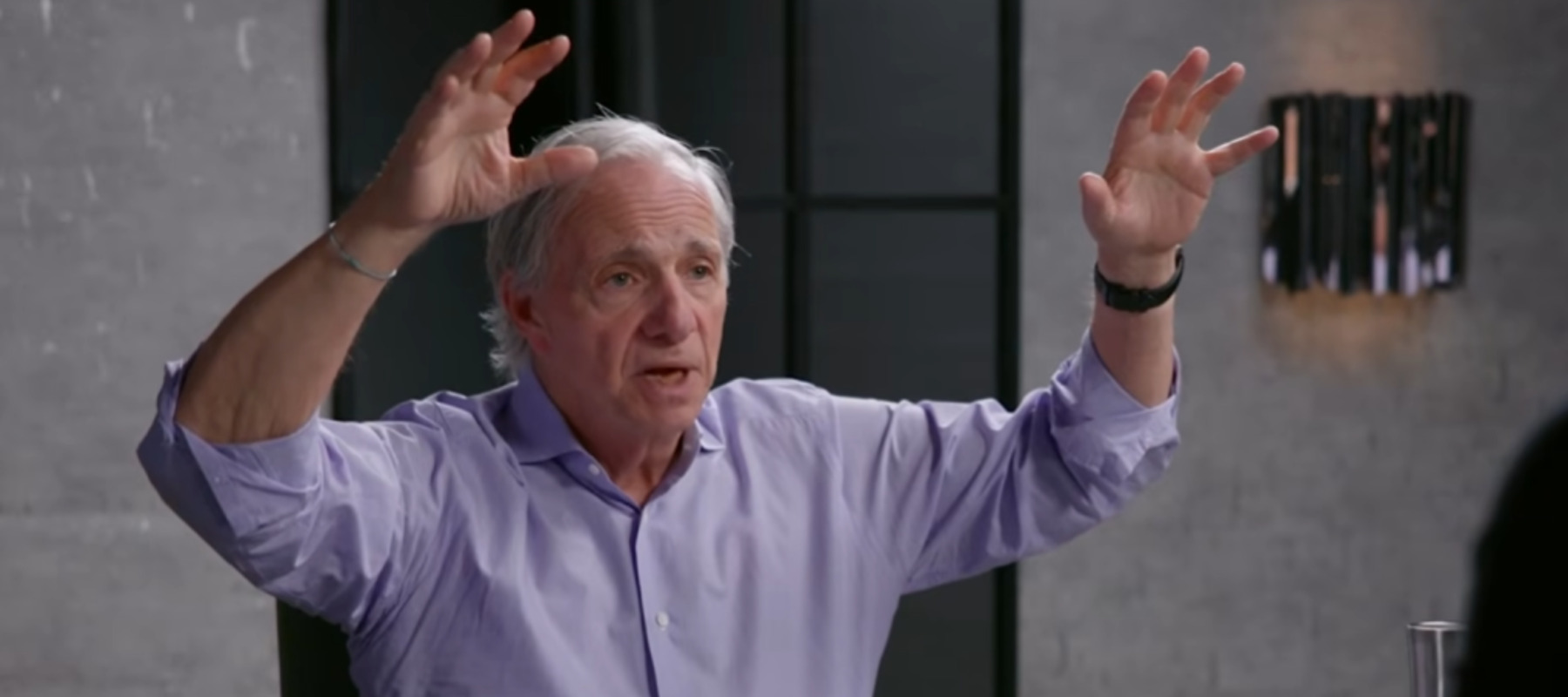Ray Dalio raises both hands as if about to surprise someone, while wearing a lavandar shirt.