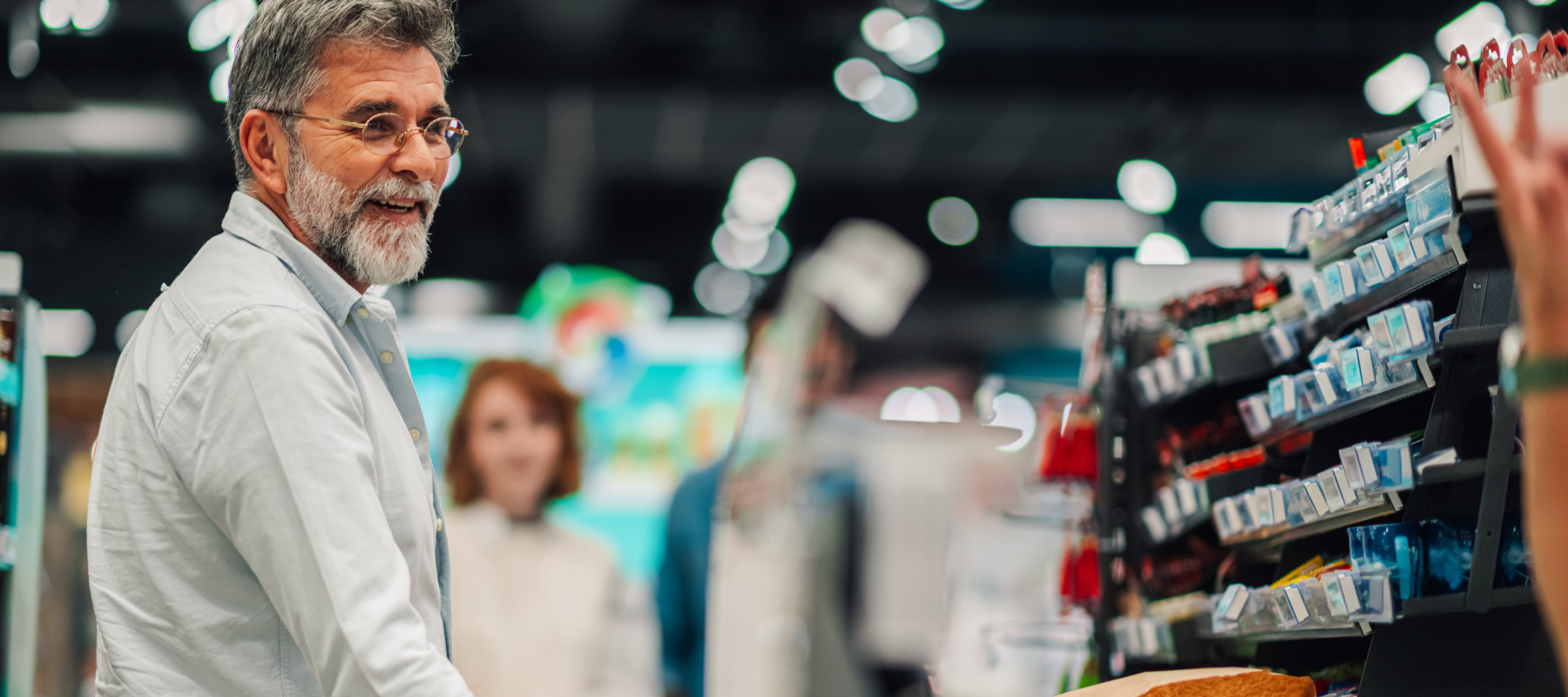 In a brightly lit grocery store, an adult man with grey hair, glasses and a trimmed beard smiles as he pushes a shopping cart filled with produce.