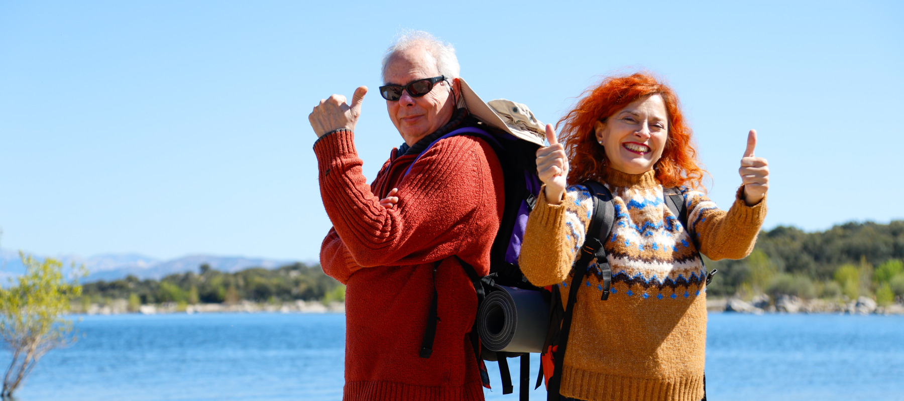 Couple in colorful clothing pose while hiking outdoors, smiling and with their thumbs up.