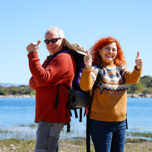 Couple in colorful clothing pose while hiking outdoors, smiling and with their thumbs up.