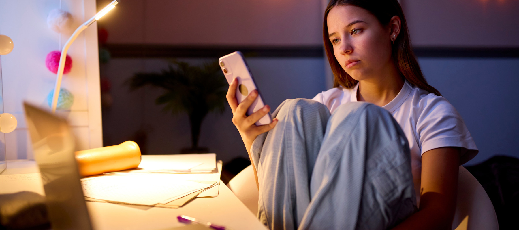 A young girl sits at her desk in her room doomscrolling on her cell phone.