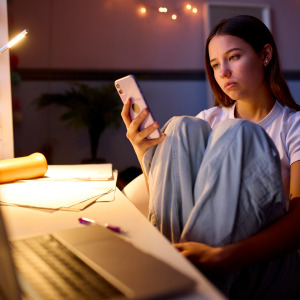 A young girl sits at her desk in her room doomscrolling on her cell phone.