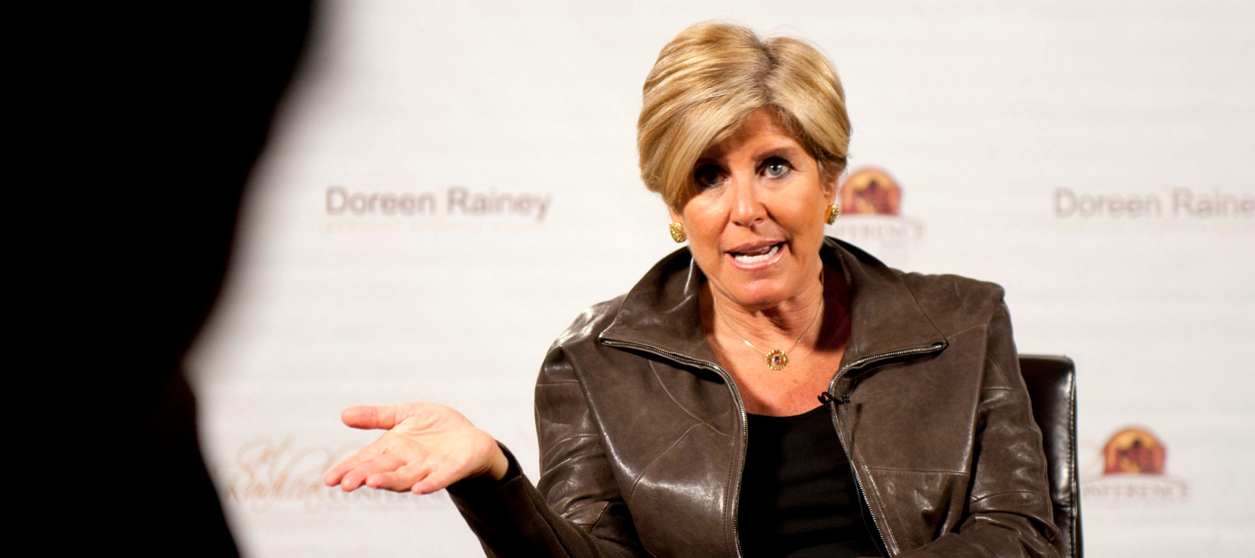 Suze Orman speaks, gesturing with her hands, while seen on stage at an event.