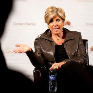 Suze Orman speaks, gesturing with her hands, while seen on stage at an event.