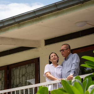Older couple seen on a balcony outside their home, talking to each other and not looking at the camera directly.