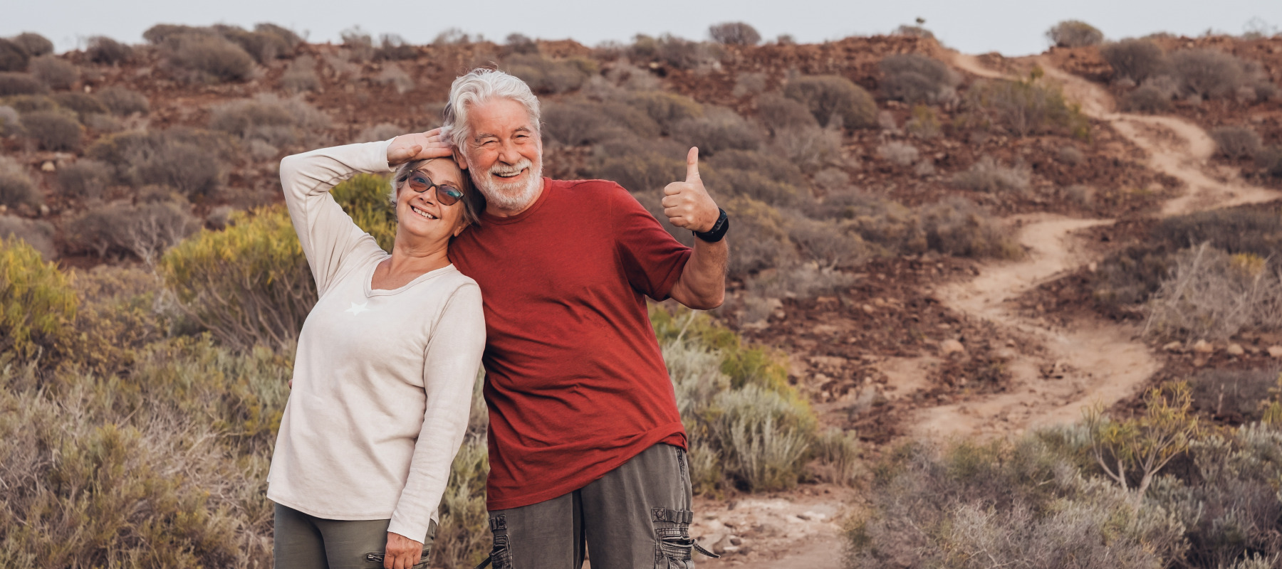 Older couple on a hike pose in front of the trail, smiling and giving a thumbs up.