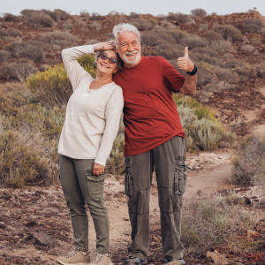 Older couple on a hike pose in front of the trail, smiling and giving a thumbs up.