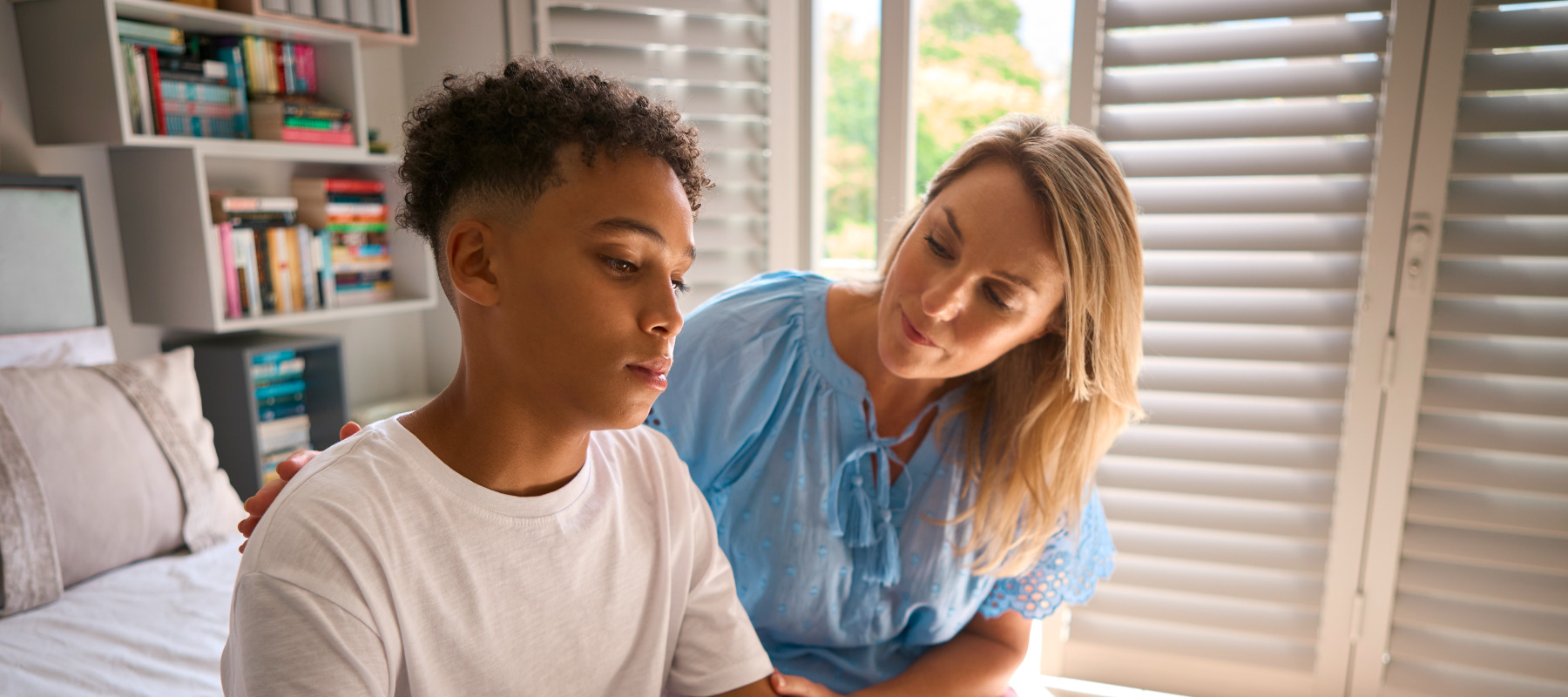 Mom comforts young teen son, who looks downcast while sitting on his bed.