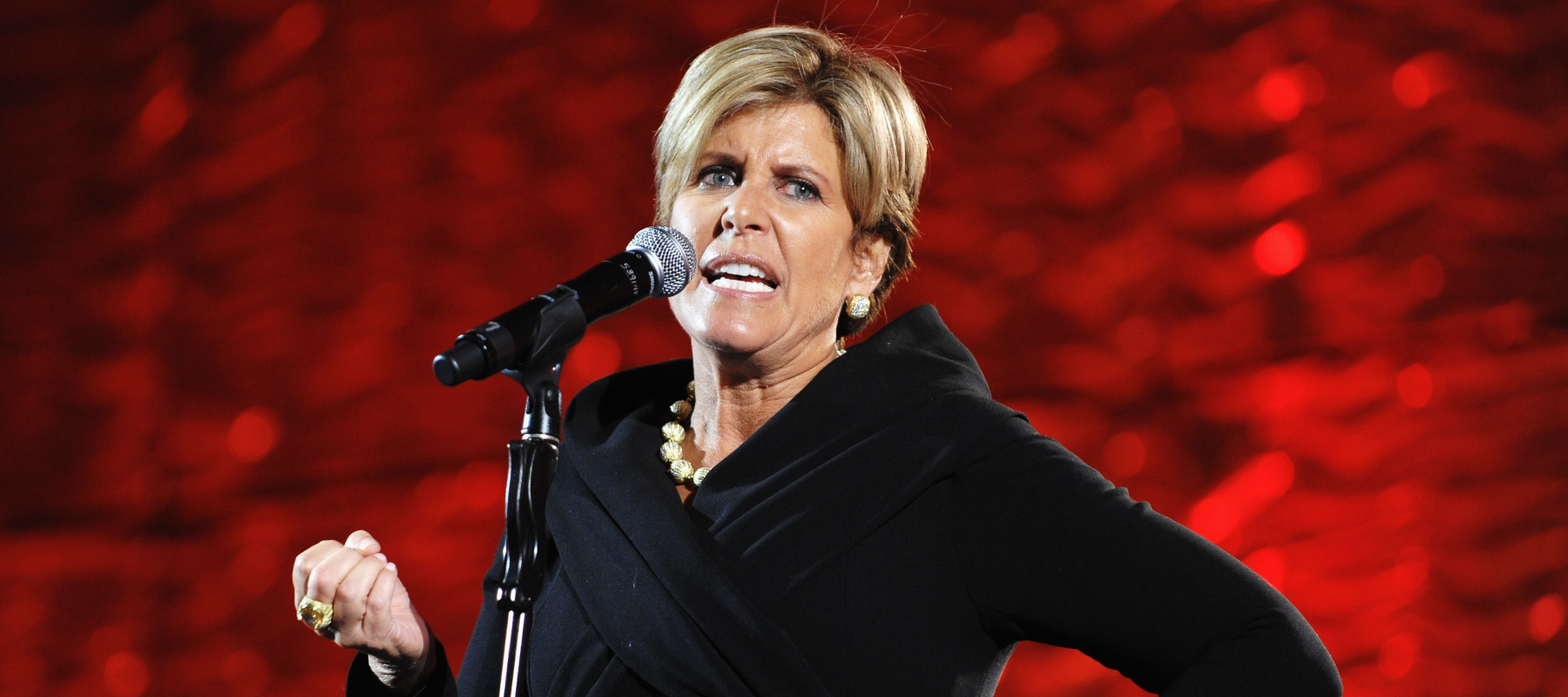 Suze Orman speaks forcefully on stage in front of a bright red backdrop.