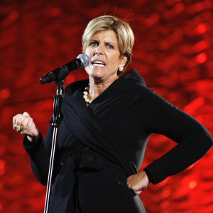 Suze Orman speaks forcefully on stage in front of a bright red backdrop.