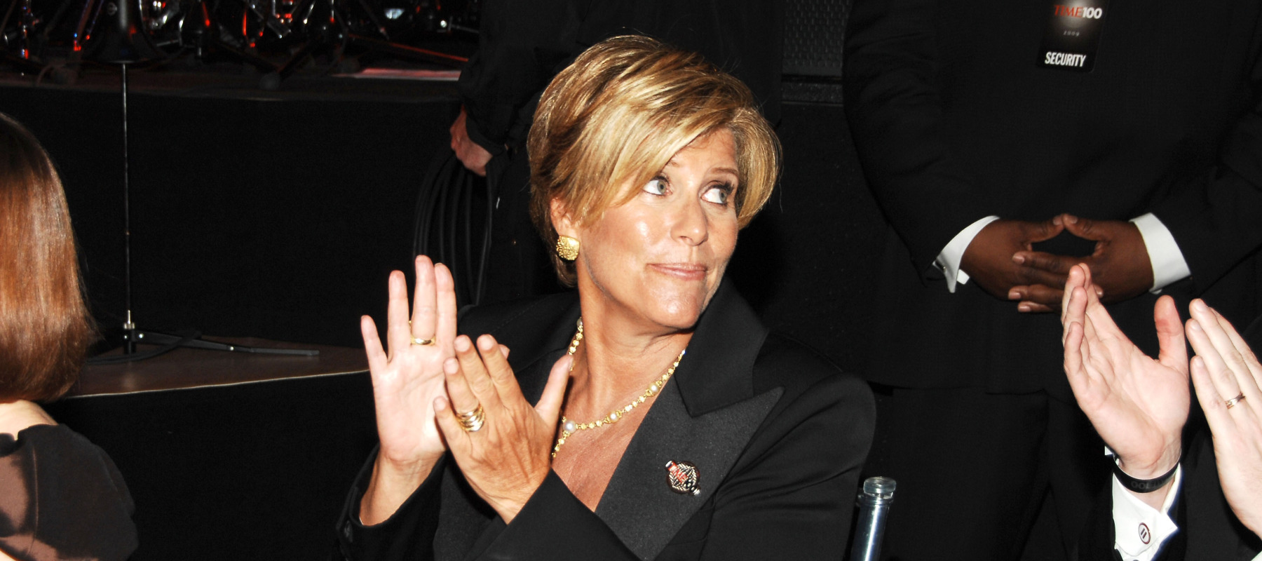 Suze Orman seen at a gala, sitting for dinner. She's clapping and looking off to the side of the camera.