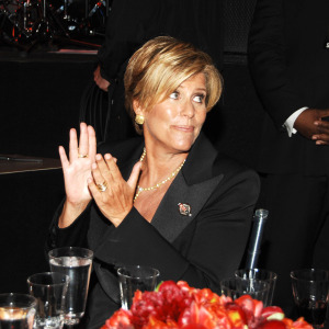 Suze Orman seen at a gala, sitting for dinner. She's clapping and looking off to the side of the camera.