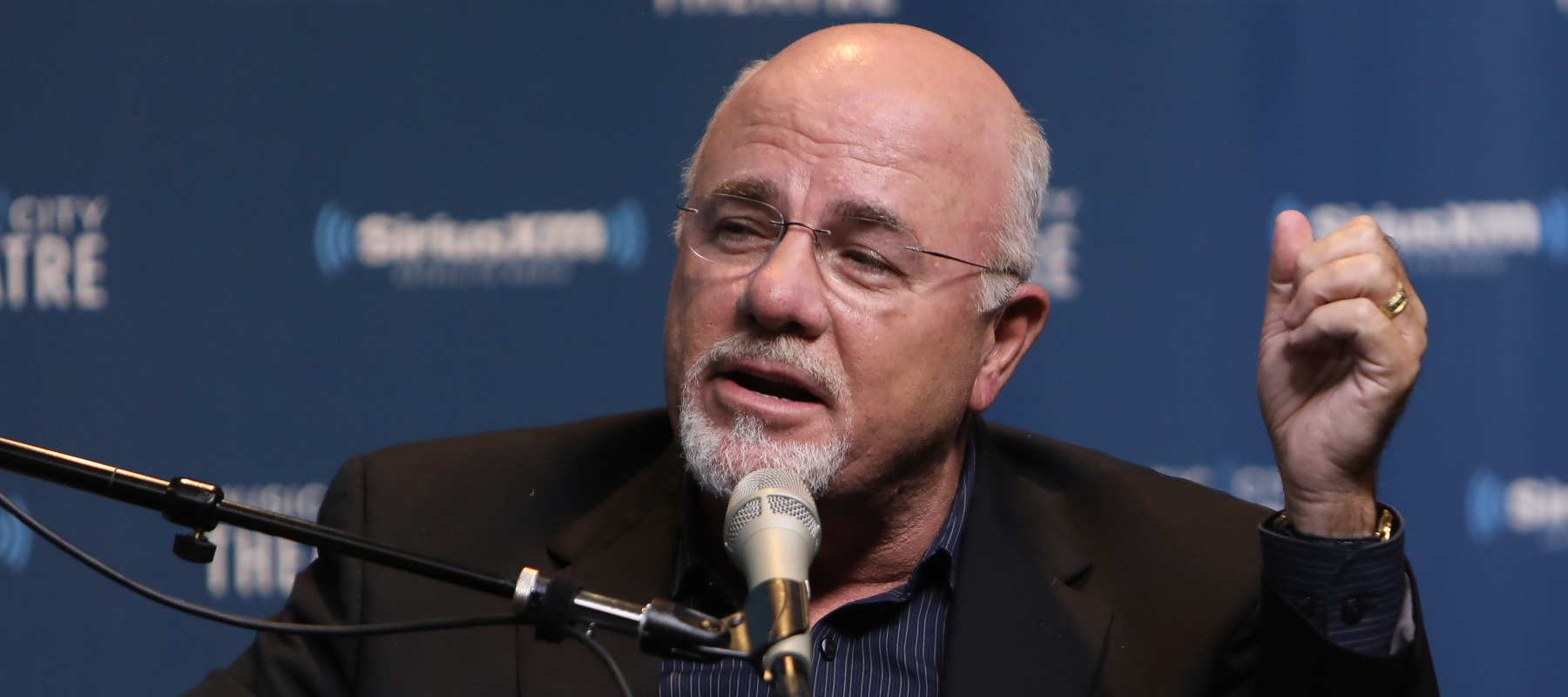 Dave Ramsey speaks into a microphone on SiriusXM while holding his fist next to his head.