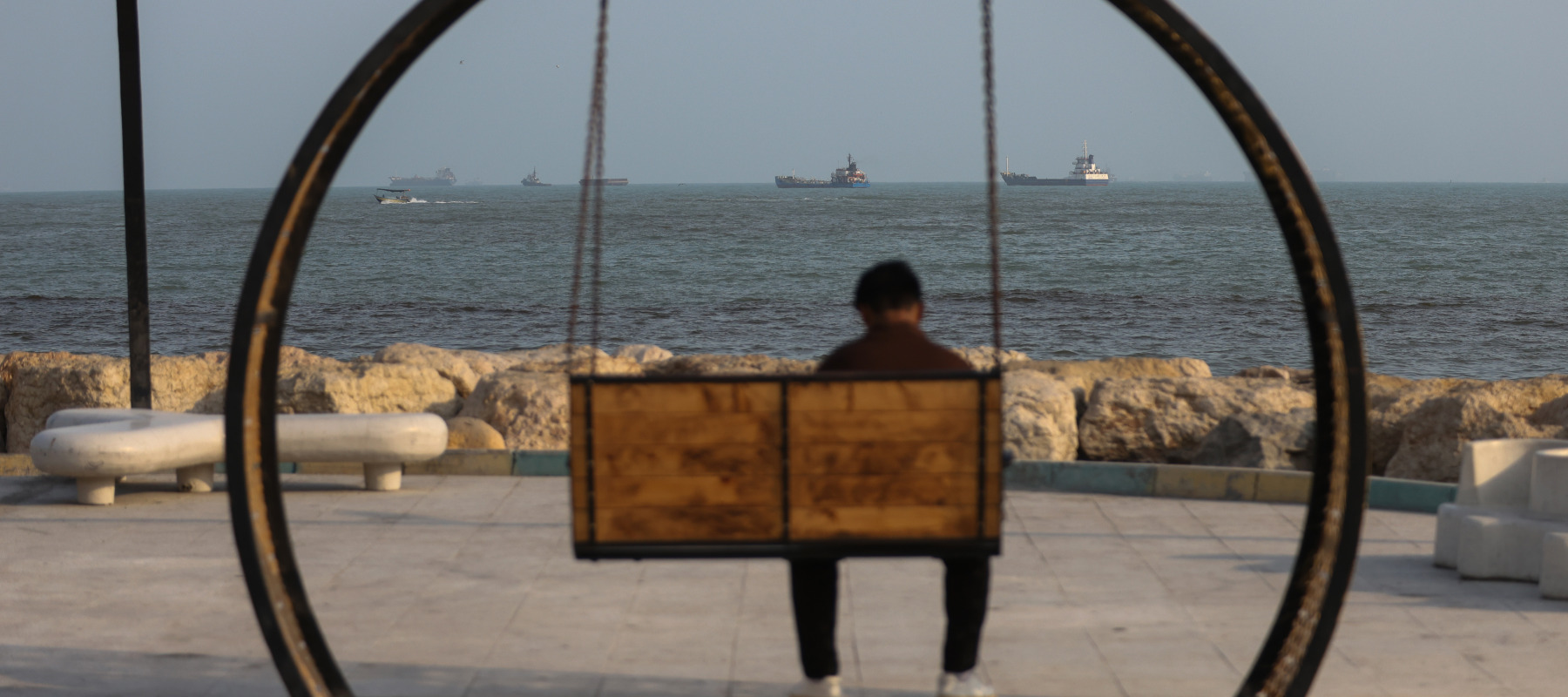 A person sits on a swing along the shoreline as ships are seen anchored in the Strait of Hormuz.