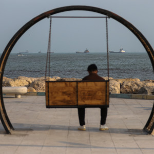A person sits on a swing along the shoreline as ships are seen anchored in the Strait of Hormuz.