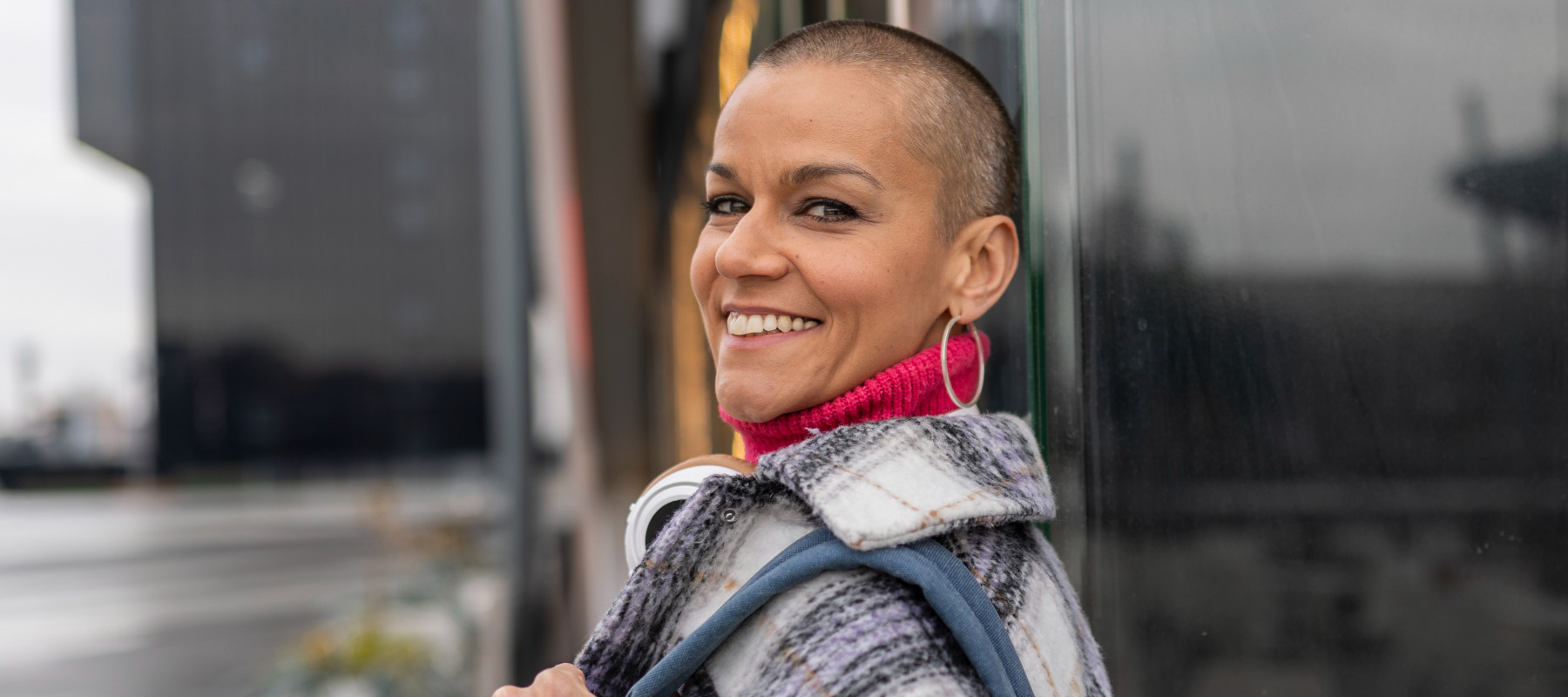 Woman with shaved head looks over her shoulder and directly at the camera, smiling.