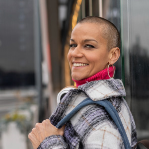 Woman with shaved head looks over her shoulder and directly at the camera, smiling.