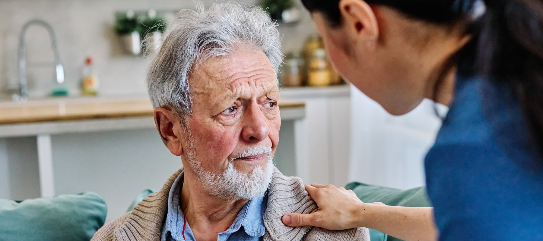 An older man in a nursing home looks away as a nurse in blue scrubs rests one hand on his shoulder.