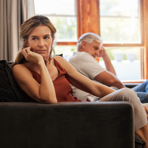 Older couple sit on the couch, looking upset and away from each other.