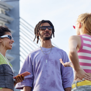 Group of Gen Z friends standing outdoors talking and laughing, all wearing sunglasses and casual outfits.