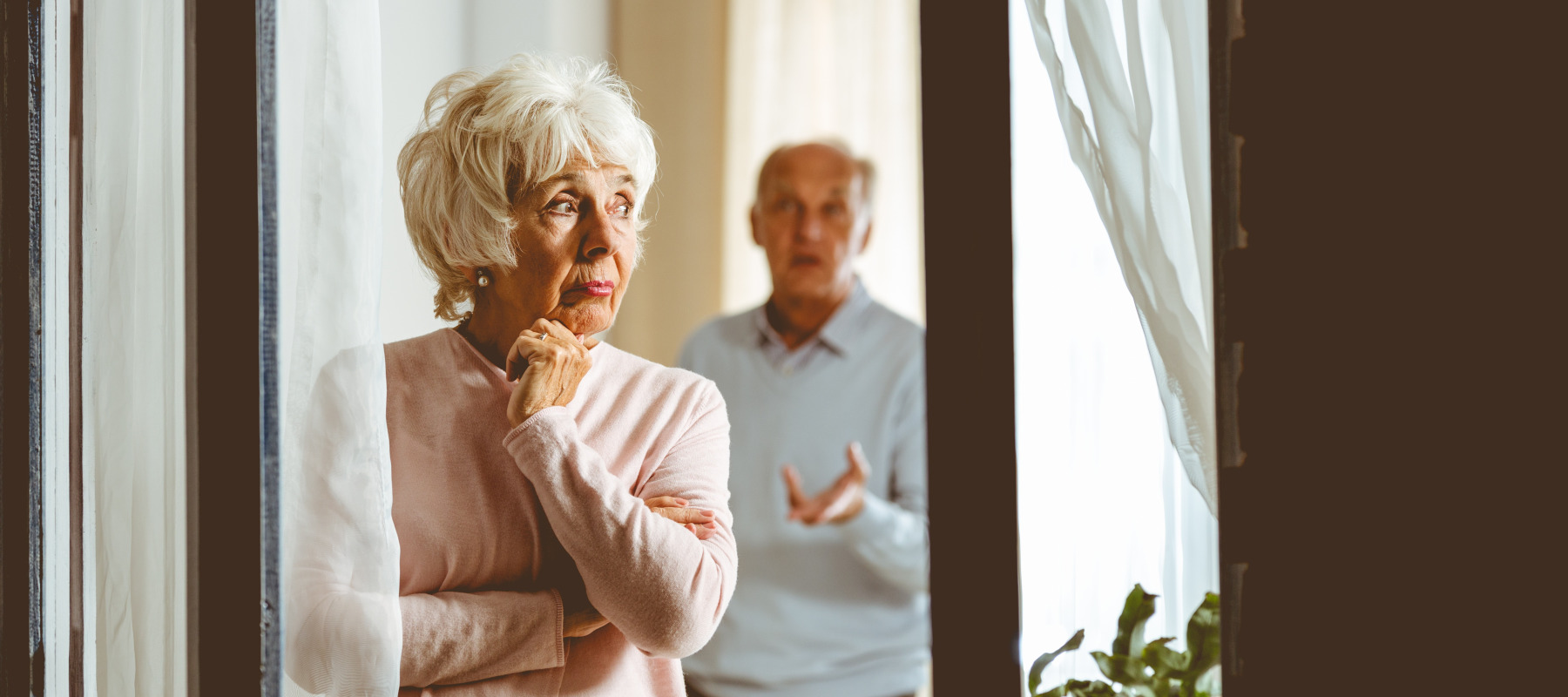 Older couple stand in front of window at home, looking uncertain and unhappy.