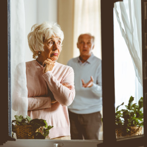 Older couple stand in front of window at home, looking uncertain and unhappy.