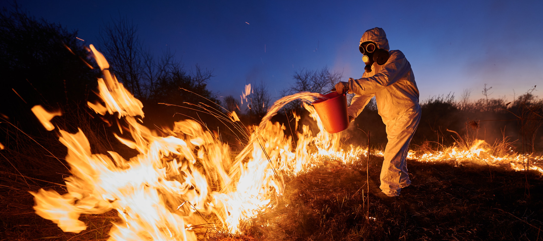 Man in a gas mask and protective suits throws bucket of water over fire.