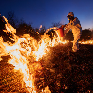 Man in a gas mask and protective suits throws bucket of water over fire.